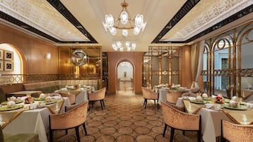 A luxurious dining area with ornate chandeliers, patterned floor tiles, and elegantly set tables with chairs.