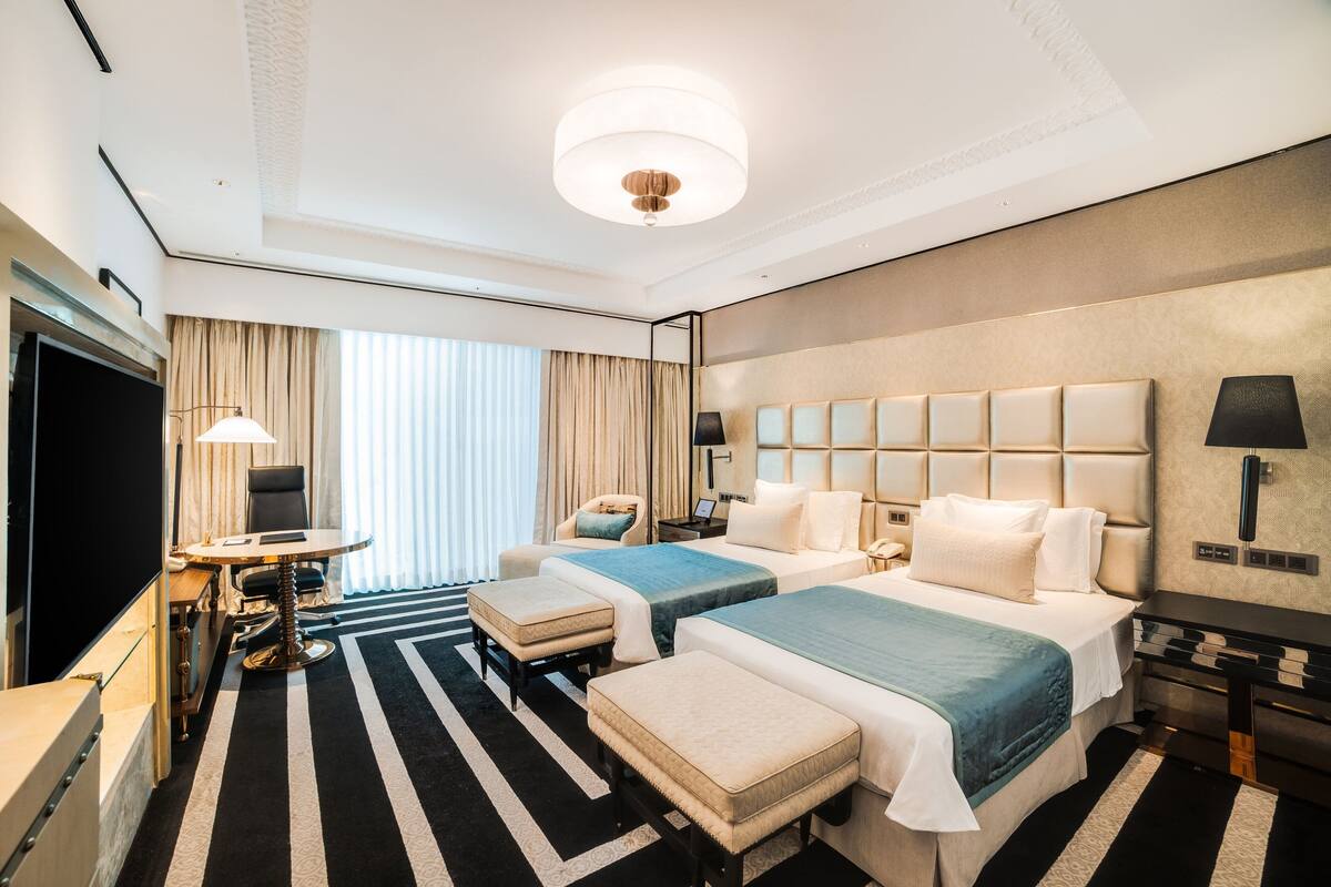 Ein Hotelzimmer mit zwei Betten, einem Fernseher, einem Schreibtisch mit Lampe und einem großen Fenster mit Vorhängen.