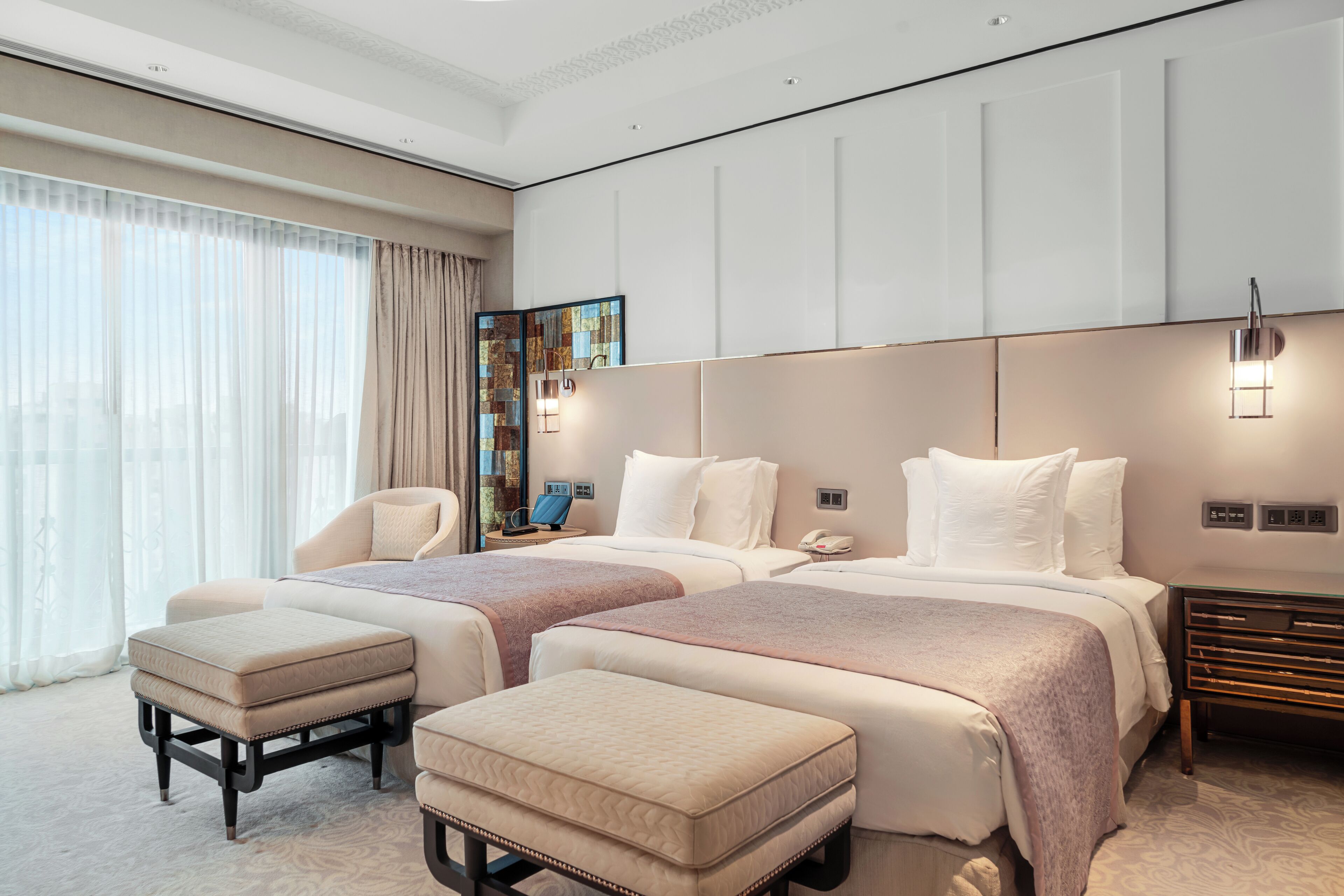 A modern hotel room with two beds, a large window with curtains, a bedside table with a lamp, and a decorative mirror.
