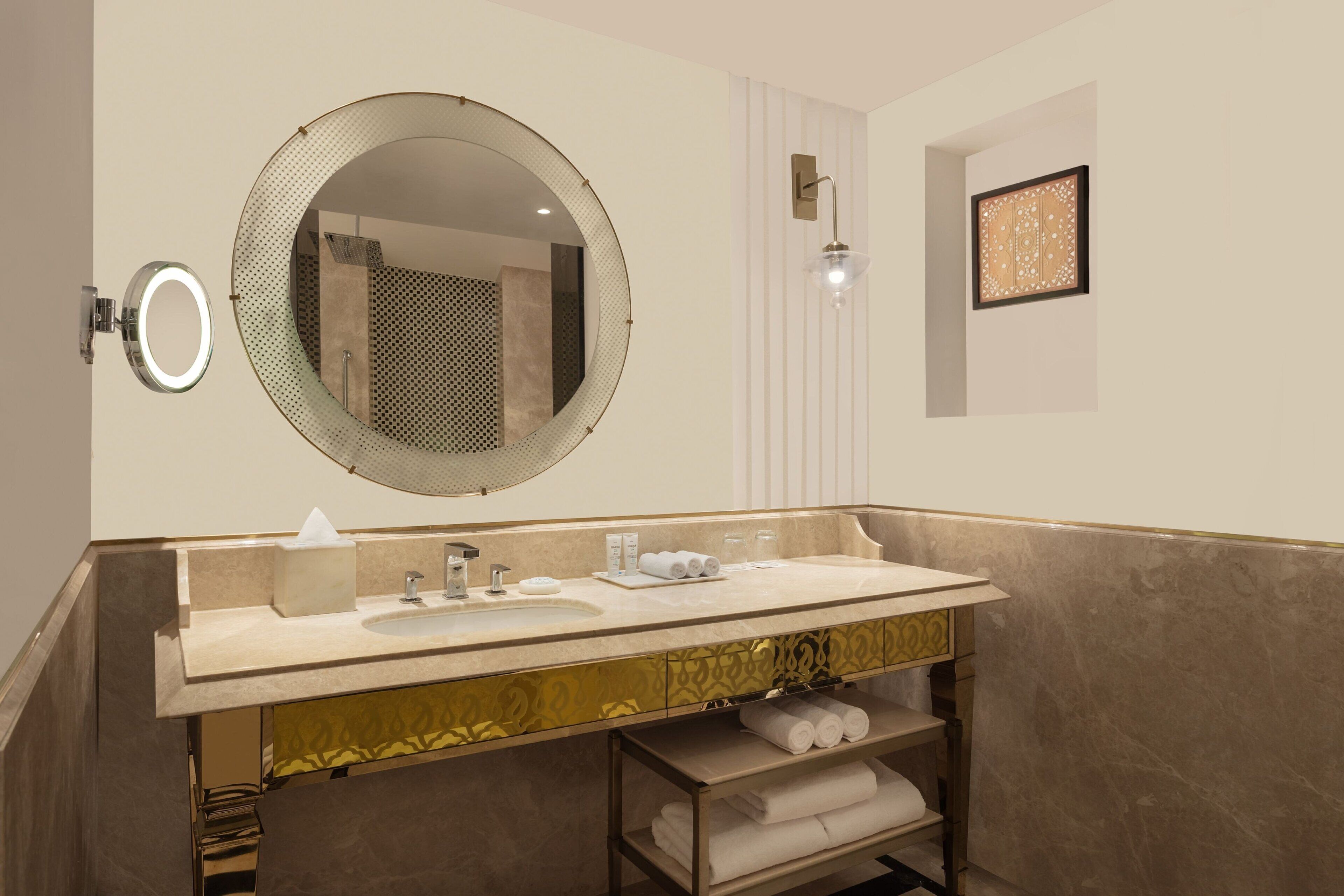 A bathroom with a round mirror, two sinks, a towel rack, and a framed artwork on the wall.