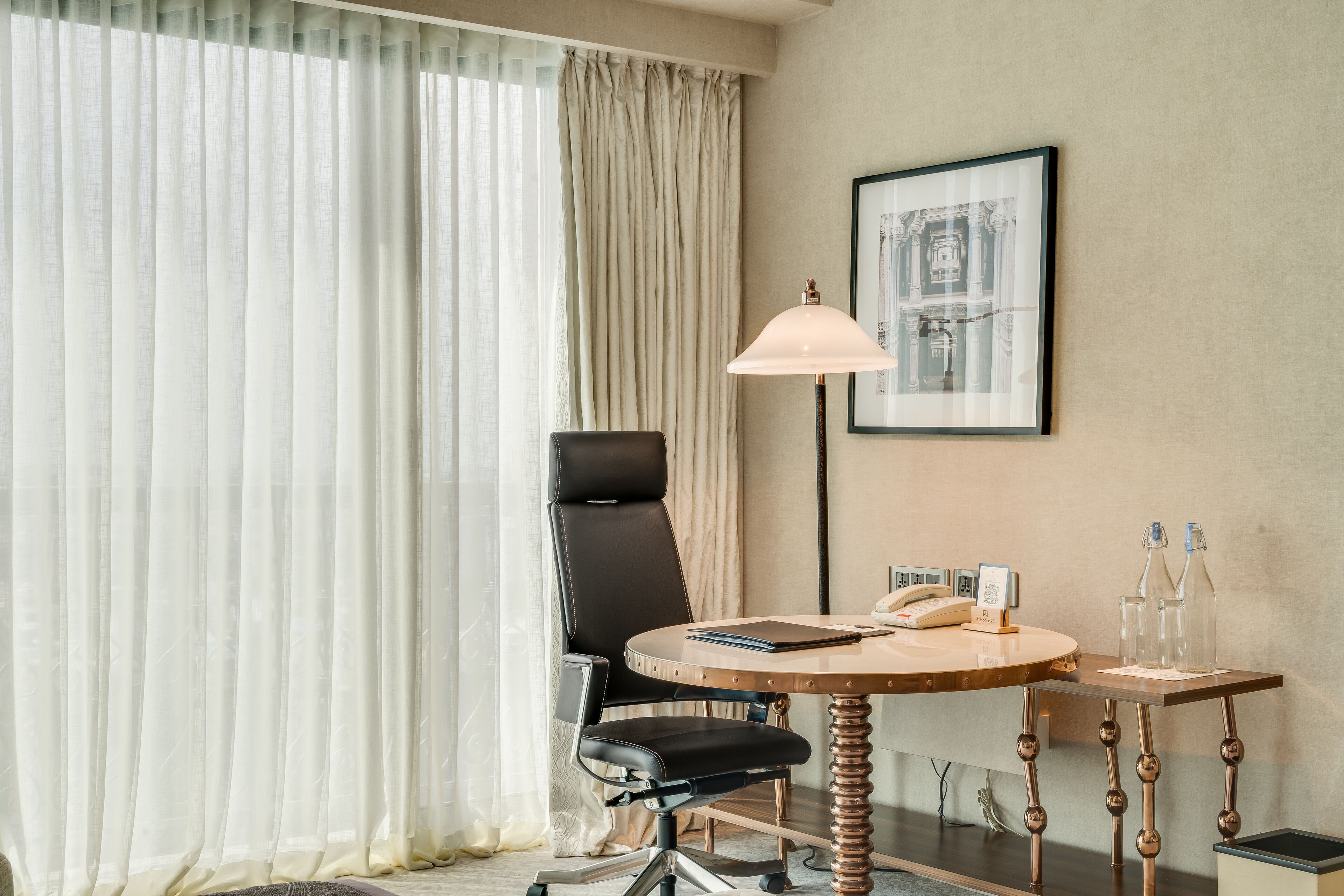Una habitación de hotel con una mesa redonda, una silla de cuero negro, una lámpara y un cuadro en la pared.