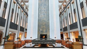 Een hotellobby met een centraal gelegen open haard, hoge plafonds en een geometrisch vloerpatroon.