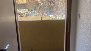 A room with a large window, a door, and a visible outdoor area with a stone wall and some trees. - Studio cabin 3 people east-facing with balcony (Valloire)