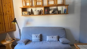 A cozy living room with a light blue sofa, two matching pillows, a wooden side table with a lamp, and a shelf above the sofa displaying various decorative items. - Studio cabin 3 people east-facing with balcony (Valloire)