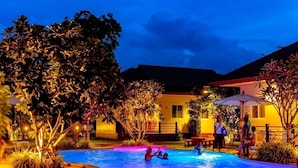 Zona de piscina por la noche con gente alrededor, rodeada de árboles y edificios.