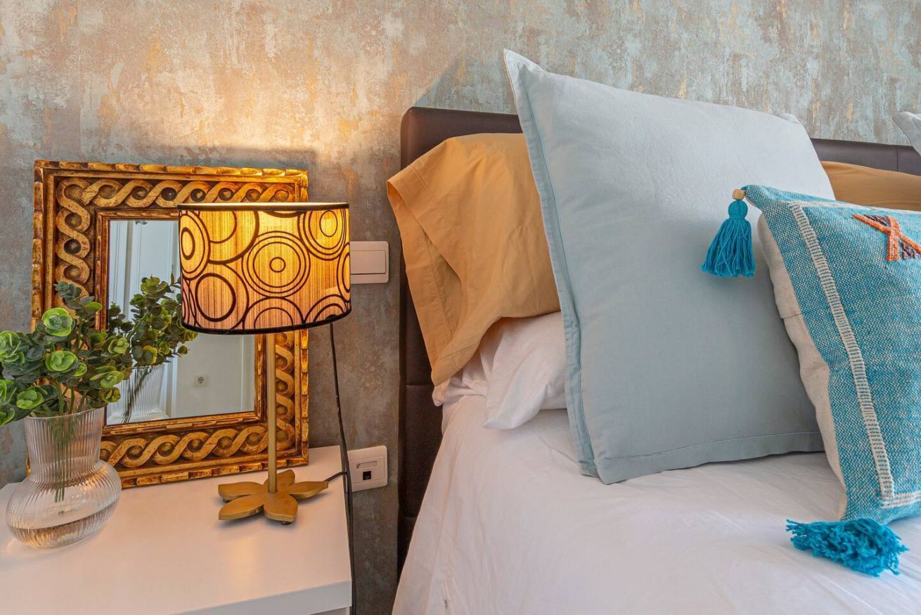 A neatly arranged hotel room with a bed, a bedside table with a lamp, a mirror, and a vase with green plants.