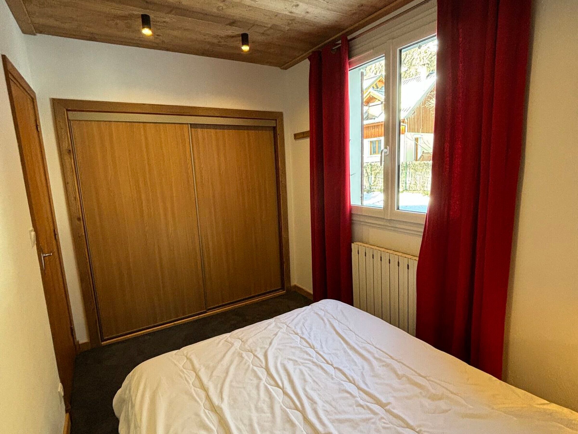A bedroom with a wooden wardrobe, a bed with white bedding, a window with red curtains, and a view of a house outside.