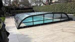 Een zwembad met glazen omheining, omgeven door een betegeld zonnedek en een haag.