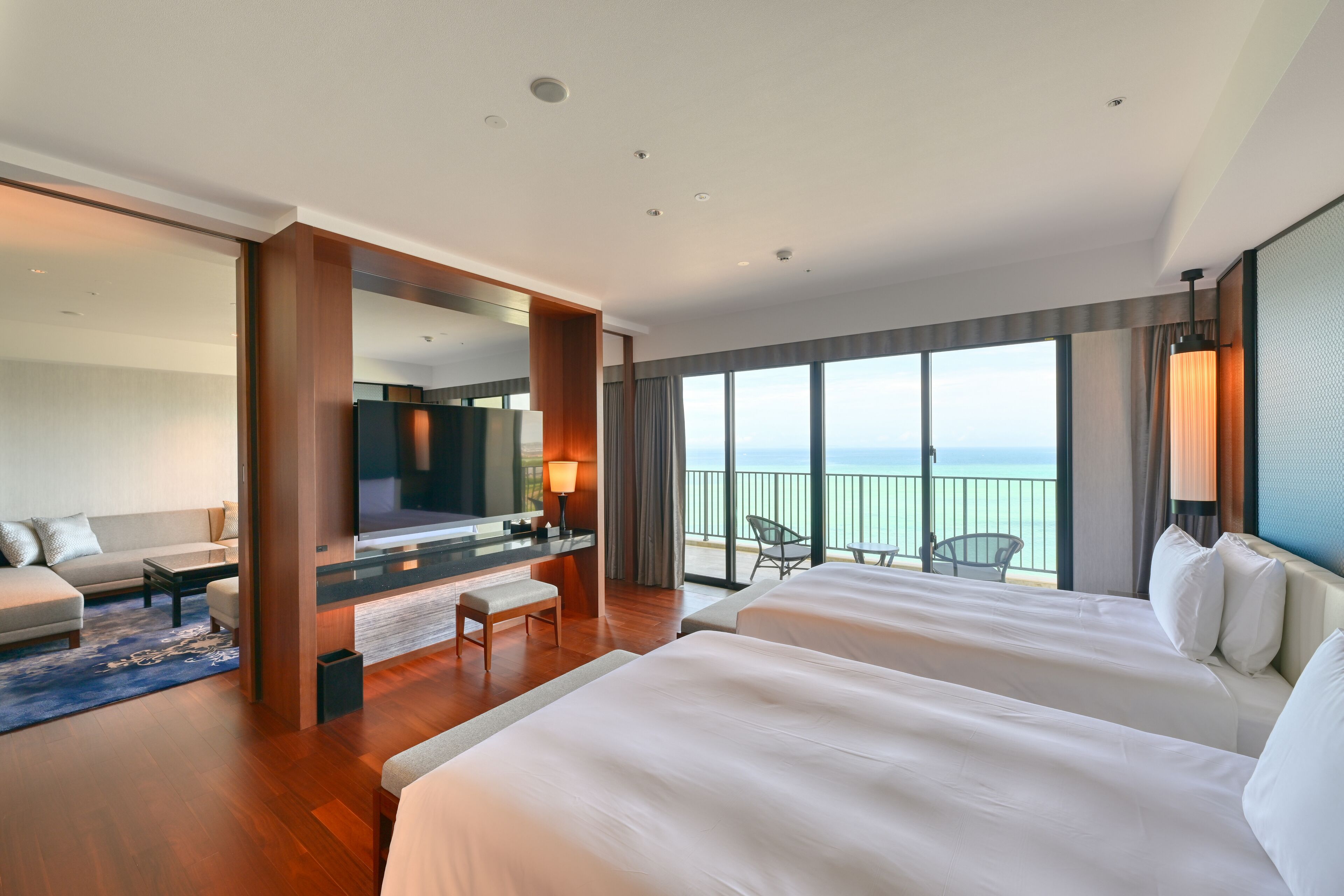 A modern hotel room with two large beds, a TV, a desk with a chair, and a balcony with a view of the sea.