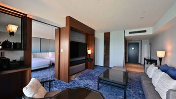 A hotel room with a bed, a TV, a coffee maker, a desk, a sofa, and a bathroom visible through an open door.
