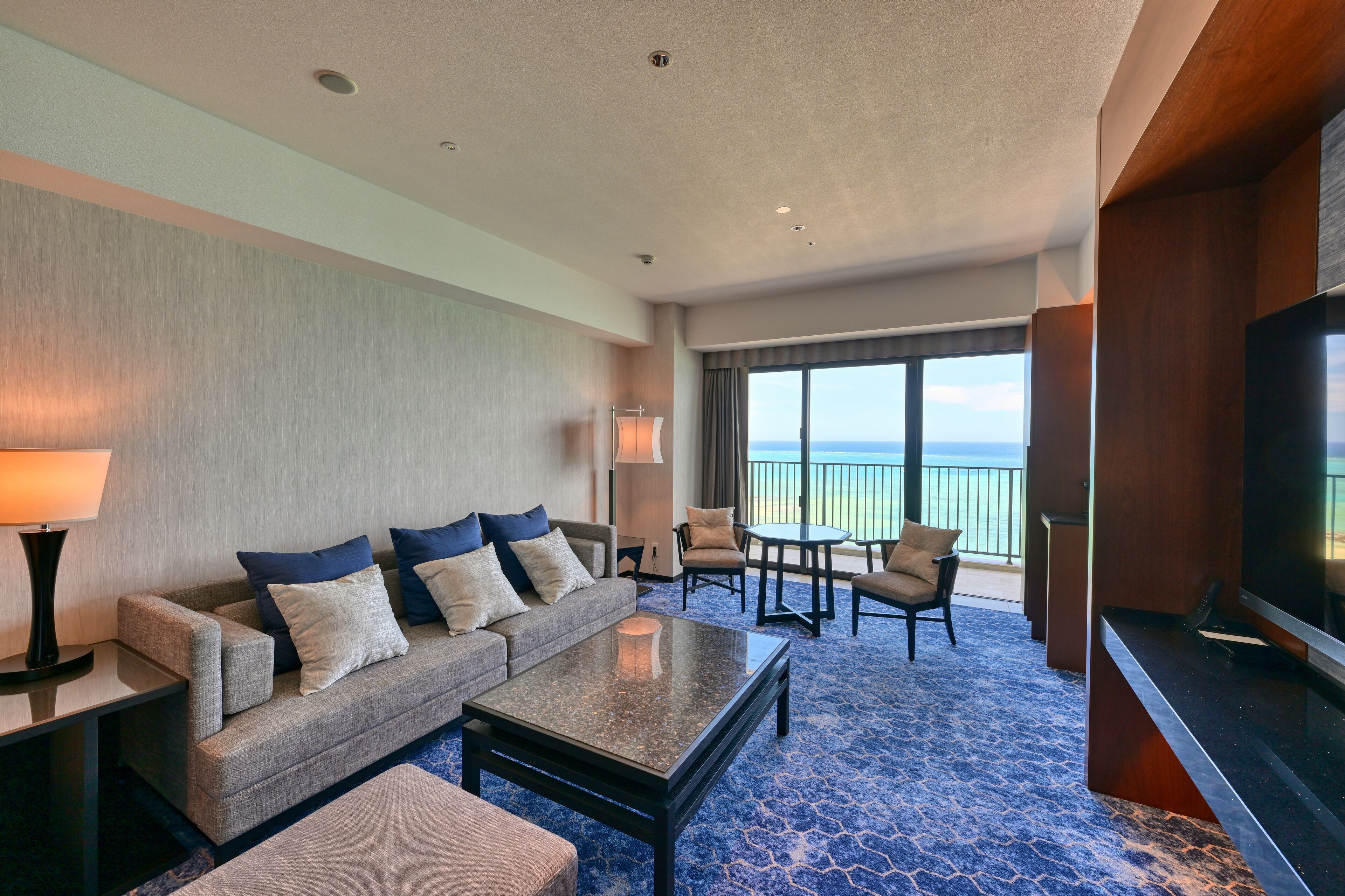 A hotel room with a sofa, armchair, coffee table, and a TV. There is a balcony with a view of the ocean.