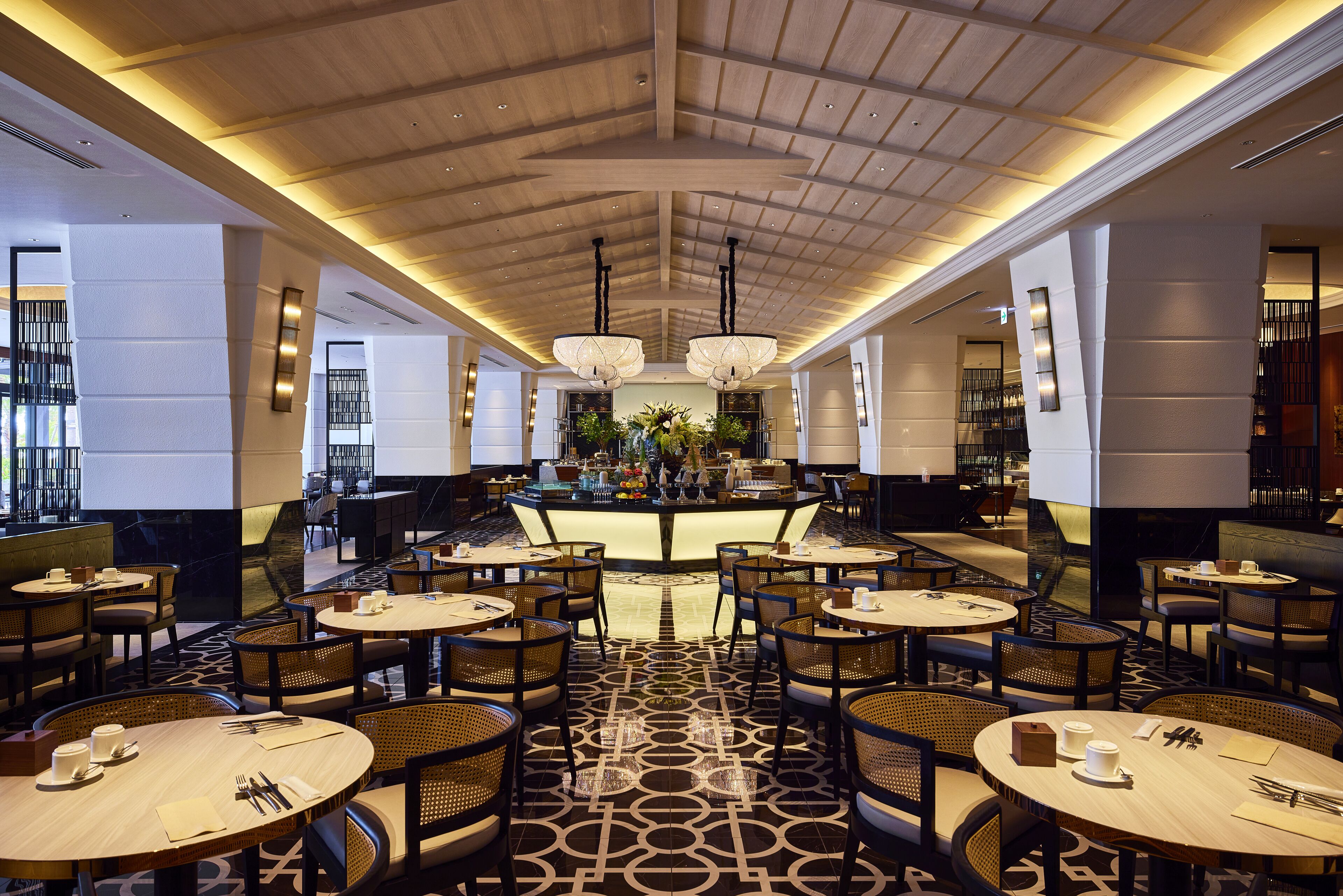 A spacious hotel lobby with round tables, comfortable seating, a central bar area, and a high ceiling with wooden beams.