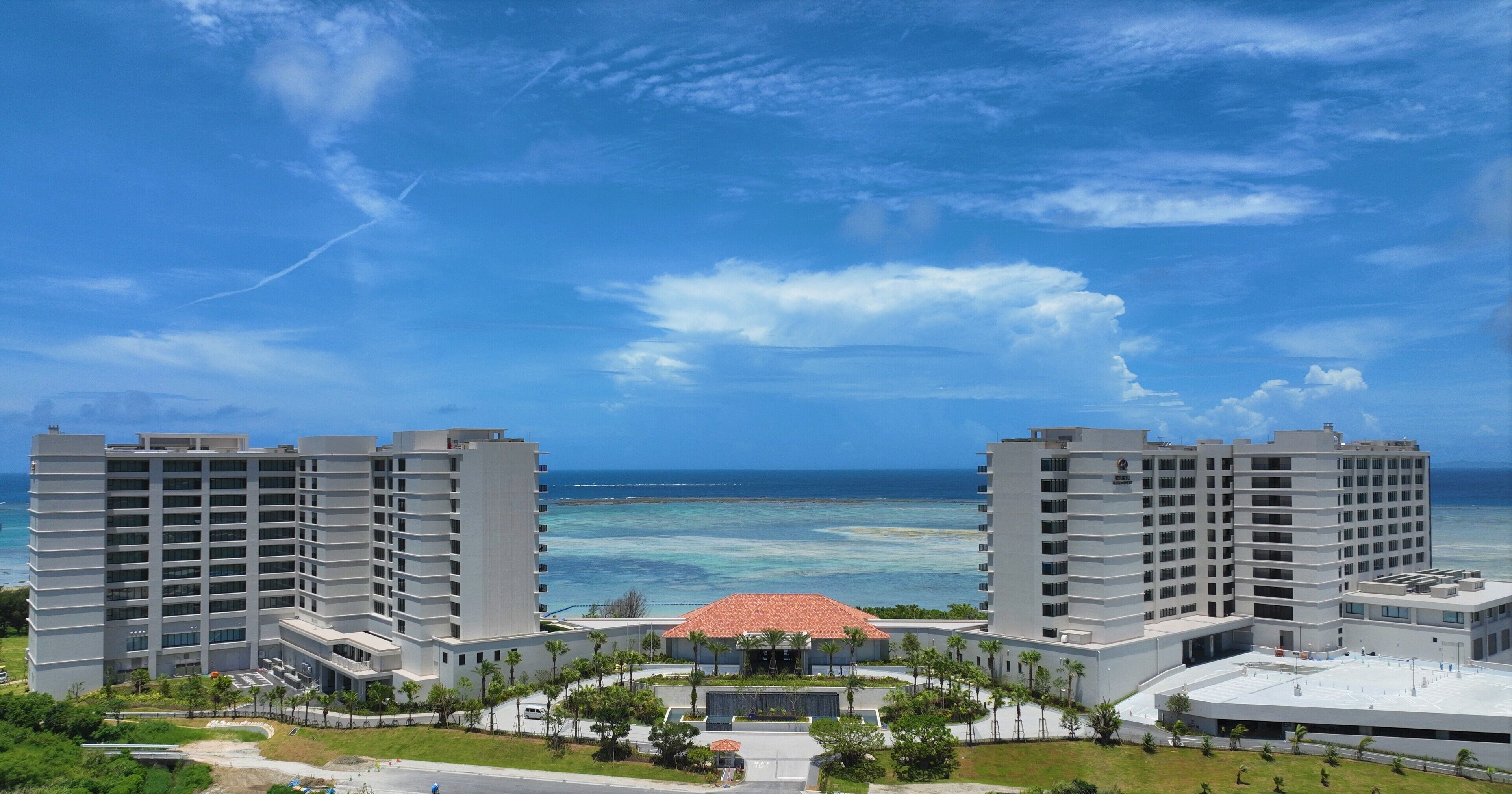 A hotel with a central building surrounded by two high-rise structures, a beach view, and a clear sky.