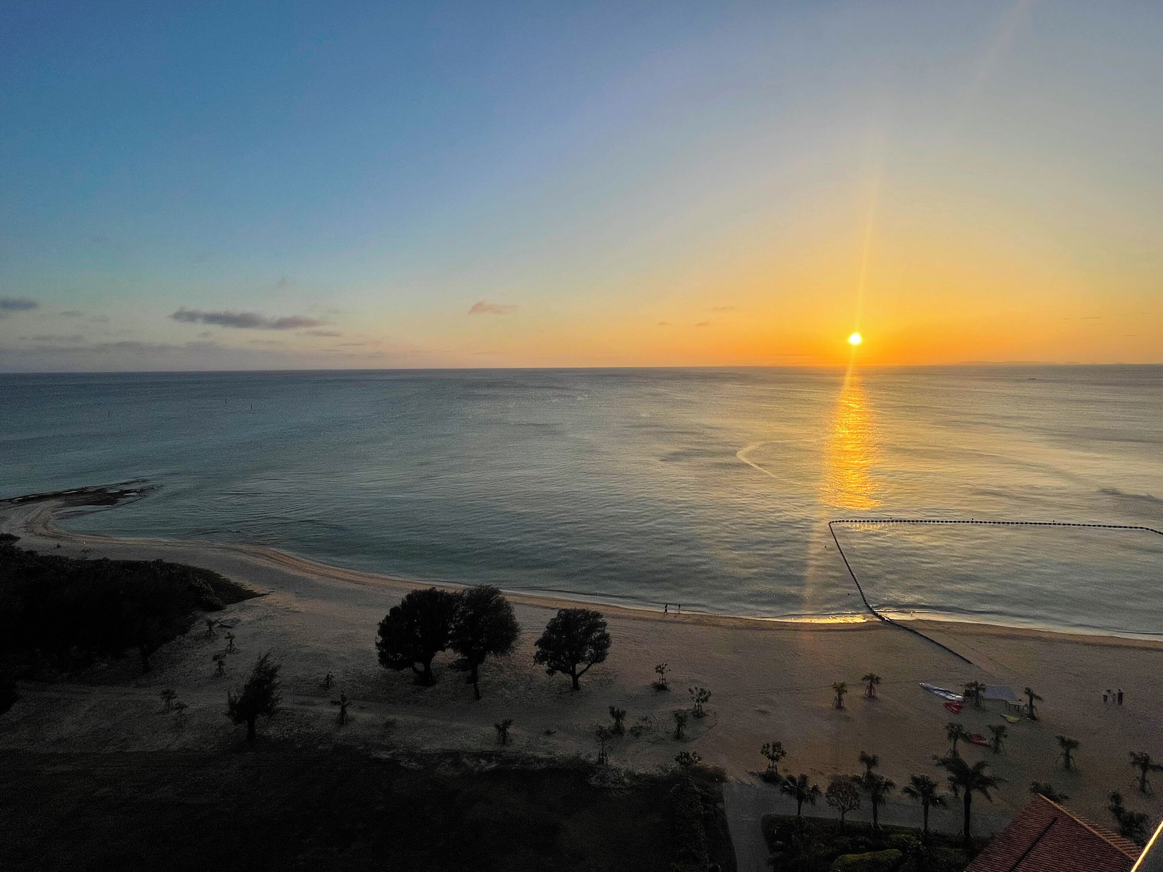 Una playa con atardecer, mar tranquilo y algunos árboles.