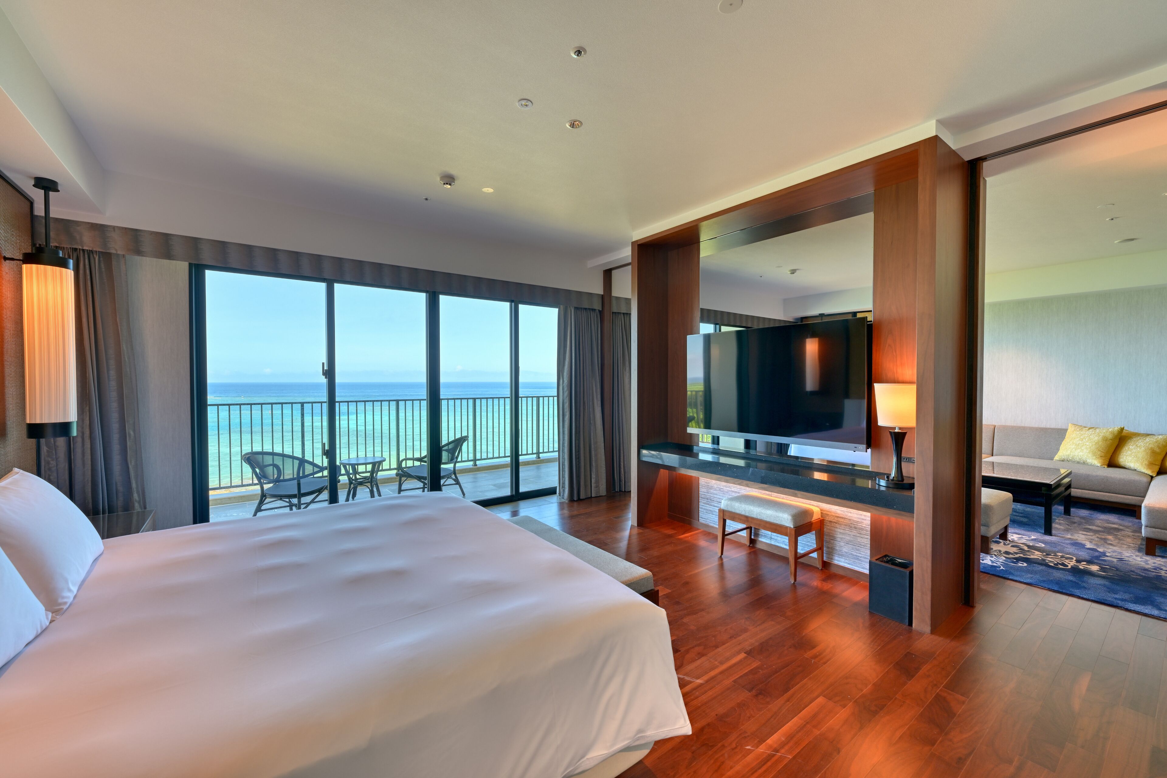 A hotel room with a large bed, a desk, a sofa, and a balcony with a view of the sea.