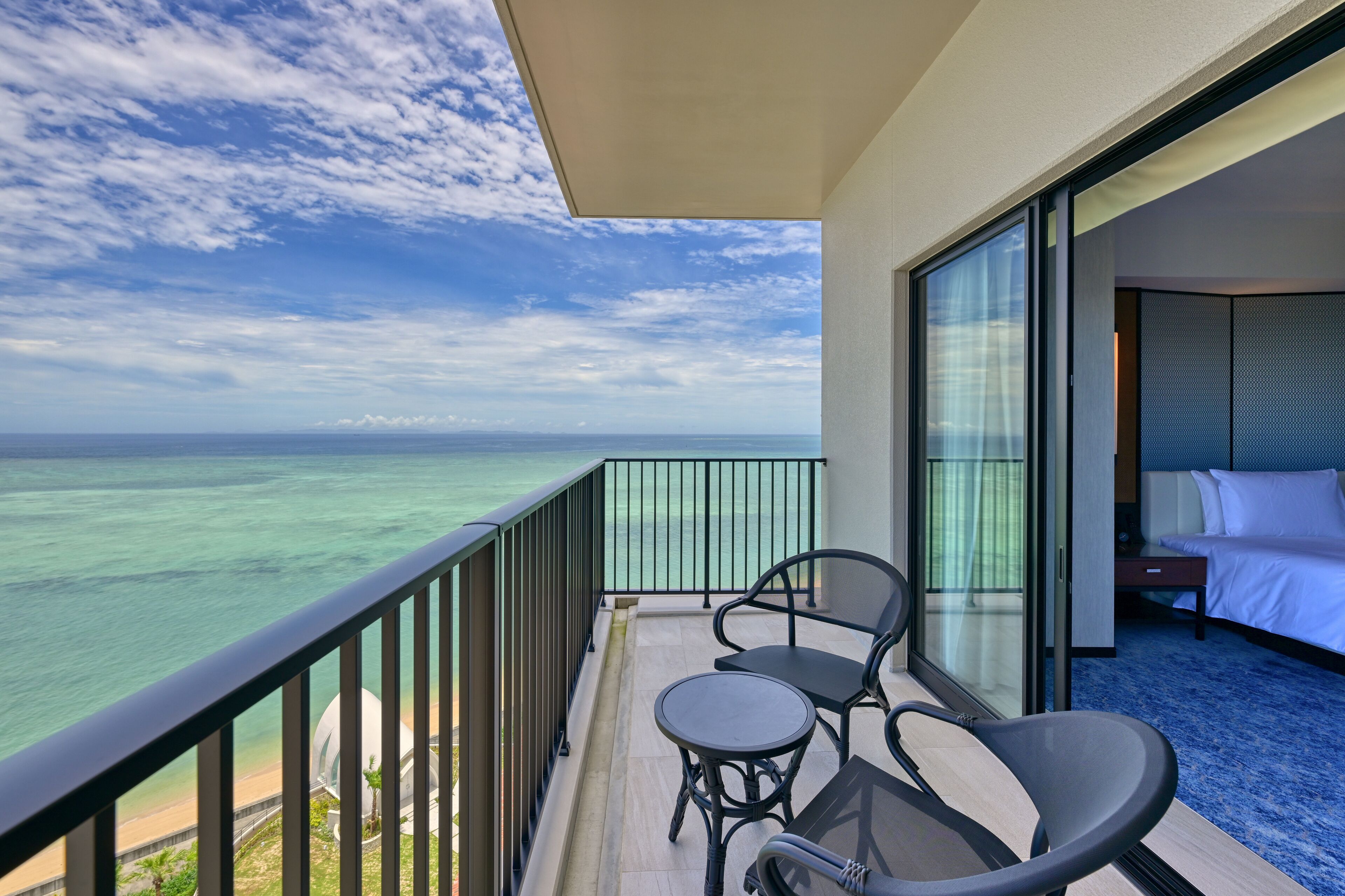 A balcony with a view of the ocean, a bed, and a small table and chair.