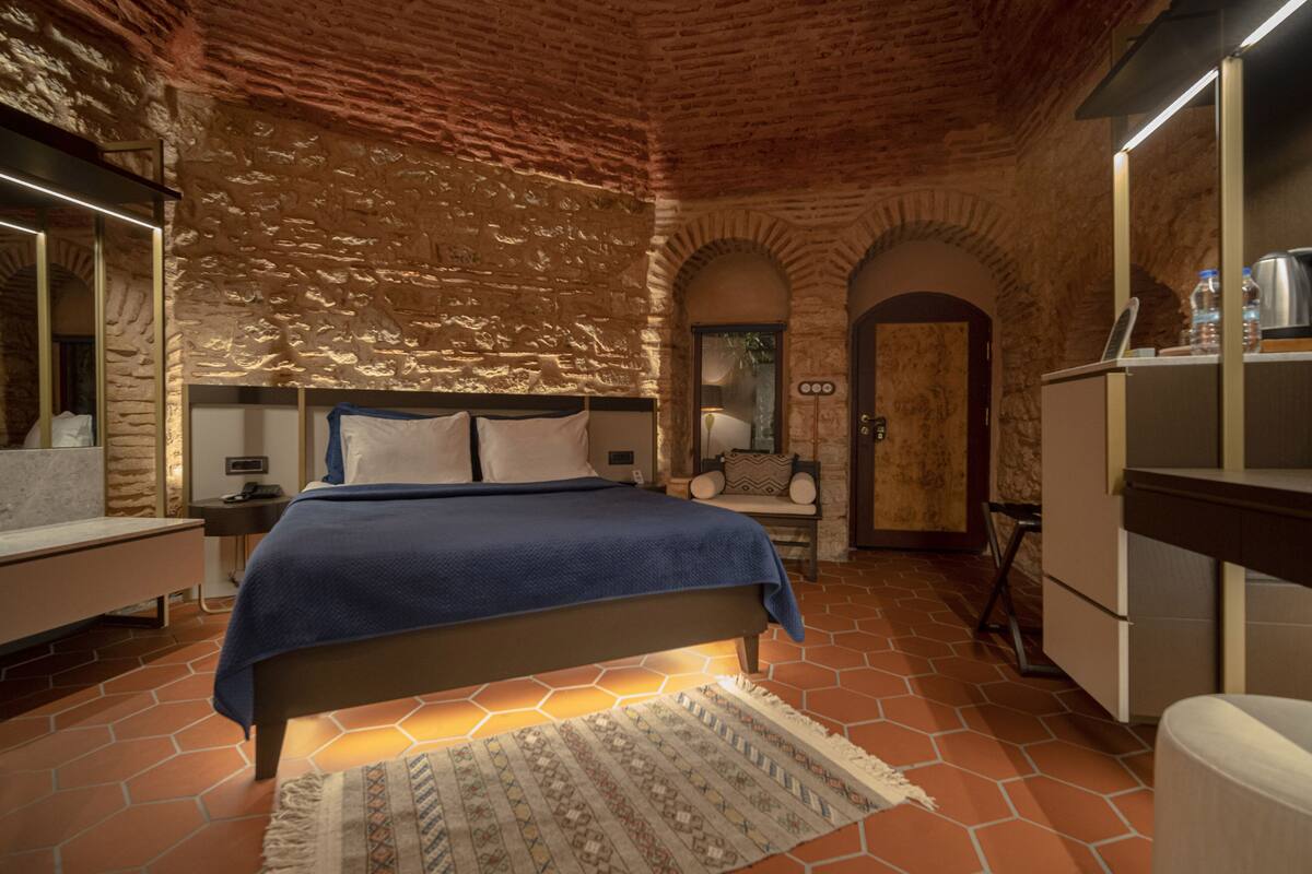 A bedroom with a stone wall, a bed with a blue blanket, a nightstand, a dresser, and a mirror.