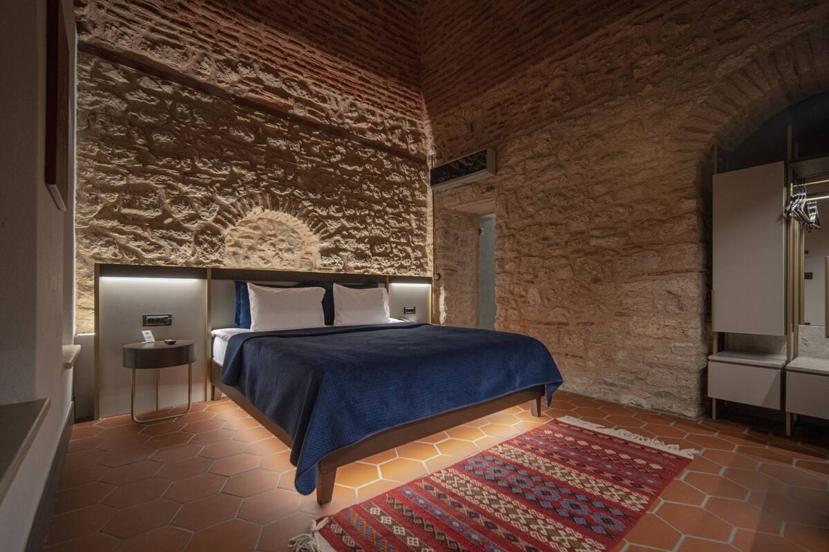 A bedroom with a stone wall, a bed with a blue blanket, a nightstand, and a red patterned rug.