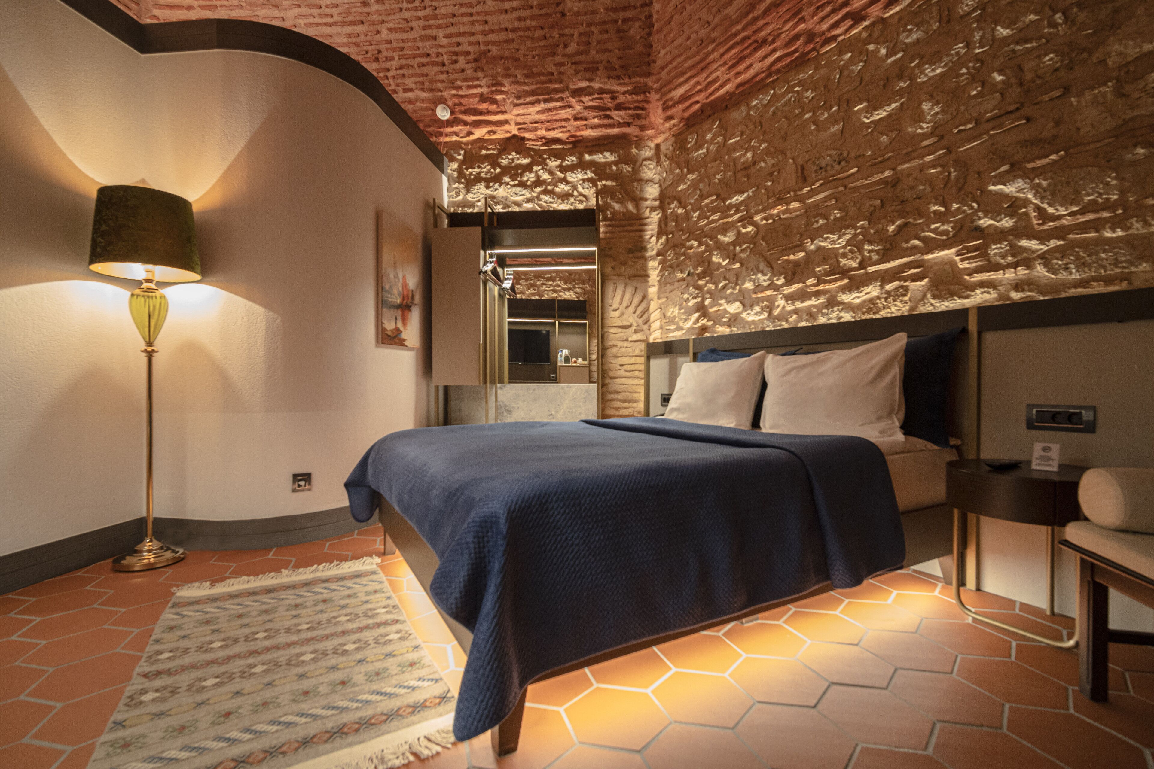 A hotel room with a brick wall, a bed with a blue bedspread, a floor lamp, and a patterned rug.