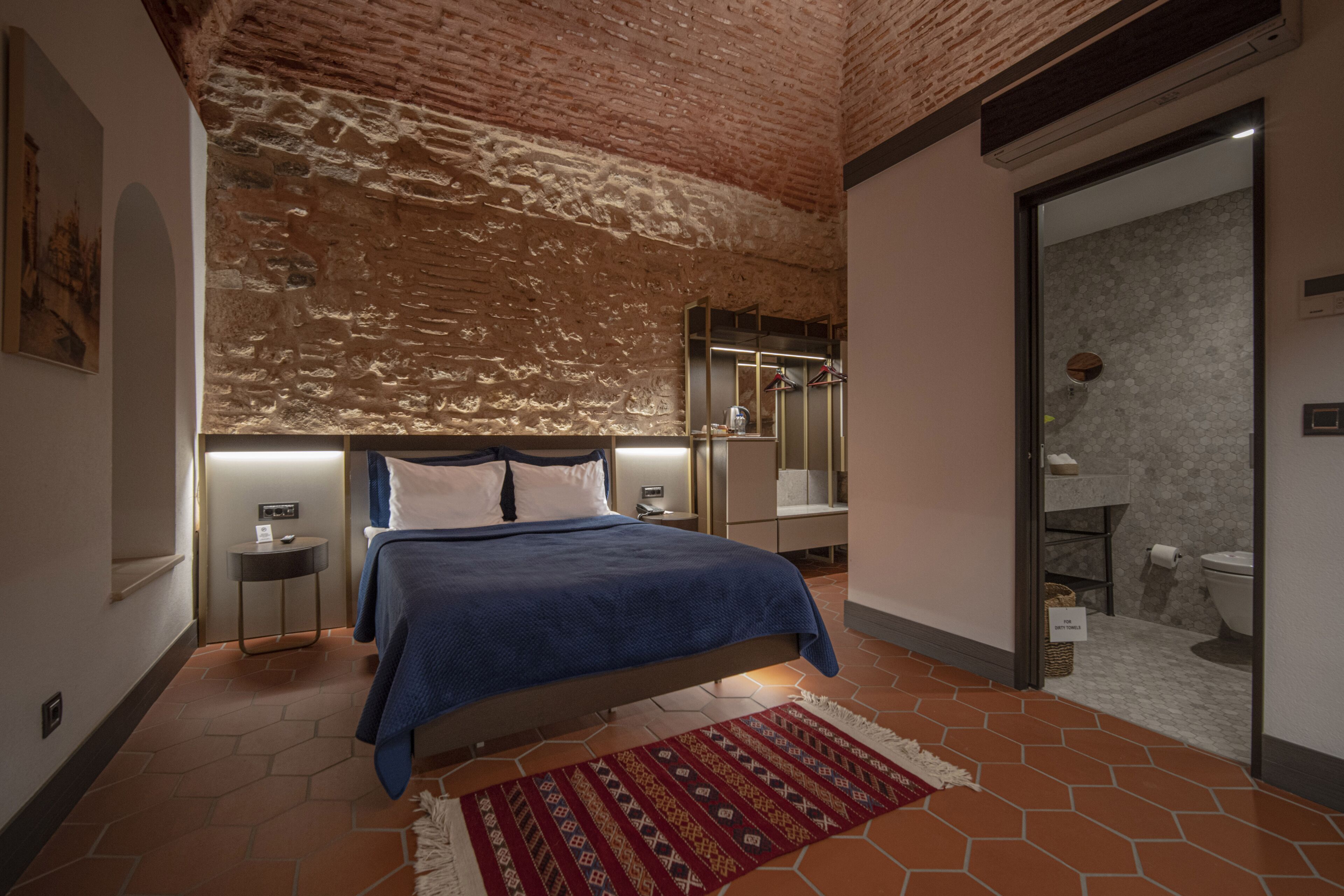 A bedroom with a brick ceiling, a bed with a blue bedspread, a nightstand, a wall-mounted shelf, and a bathroom visible through an open door.