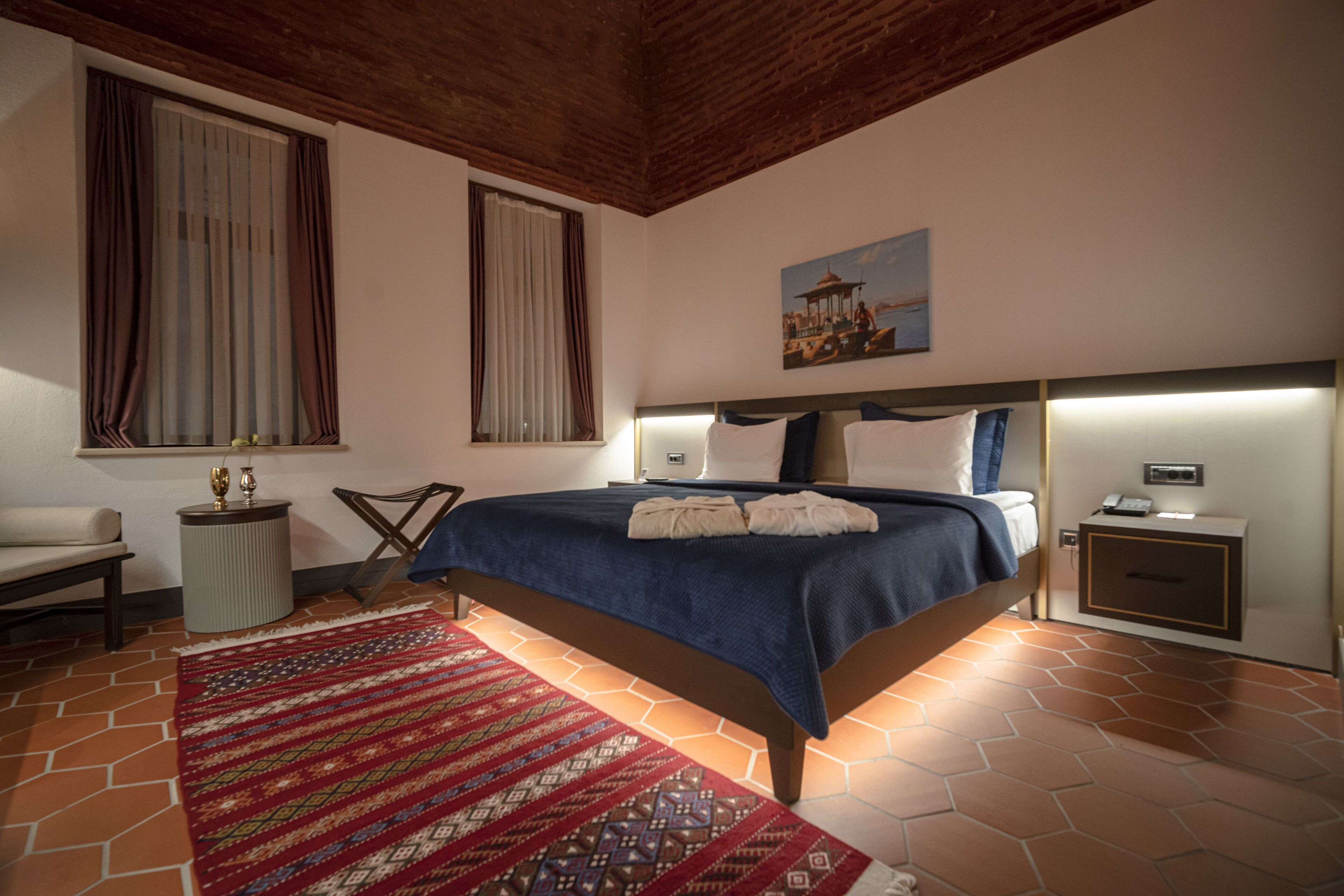 A bedroom with a bed, a nightstand, a chair, a window with curtains, and a painting on the wall.
