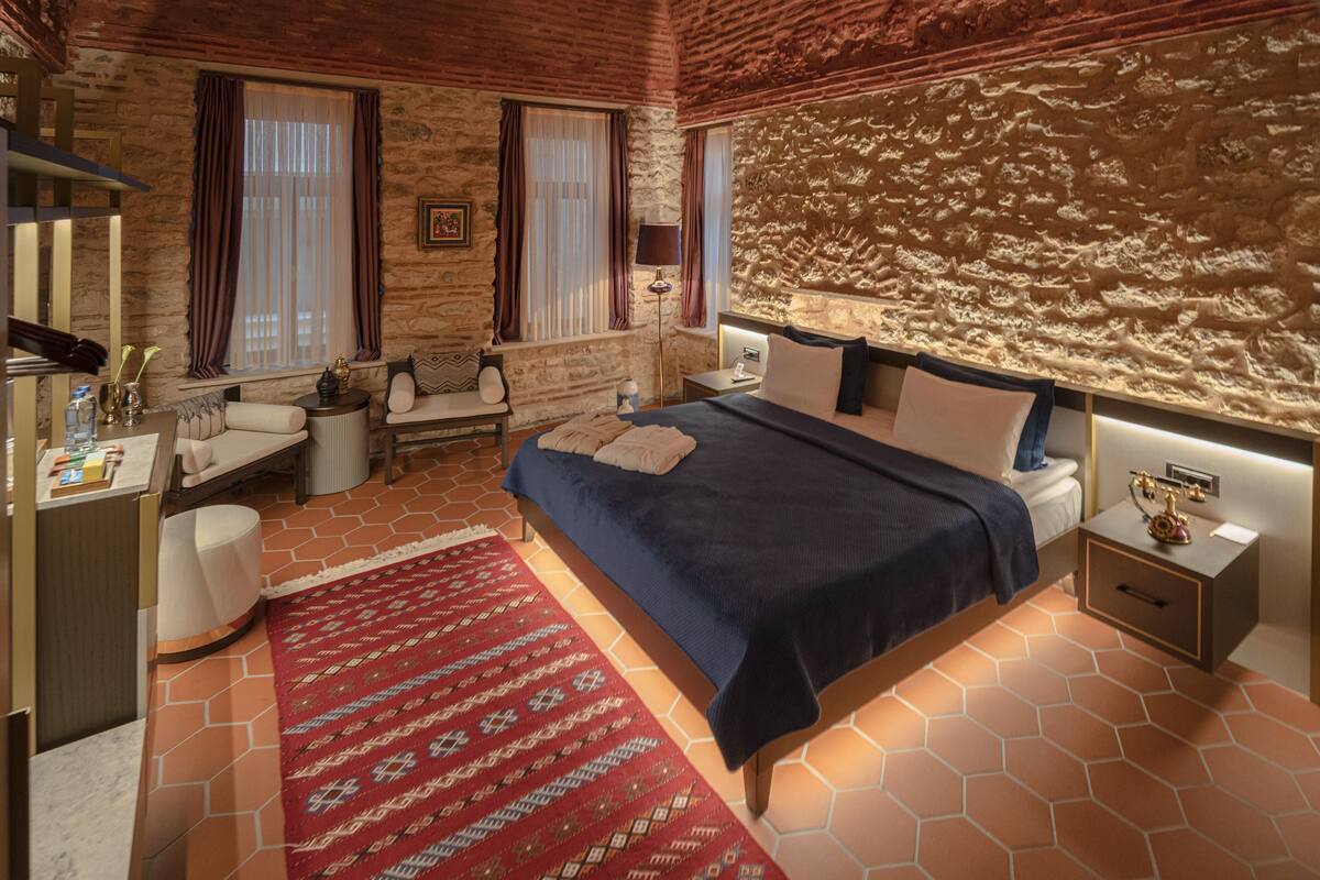A hotel room with a large bed, a desk with a chair, a small sofa, a patterned rug, and a stone wall.