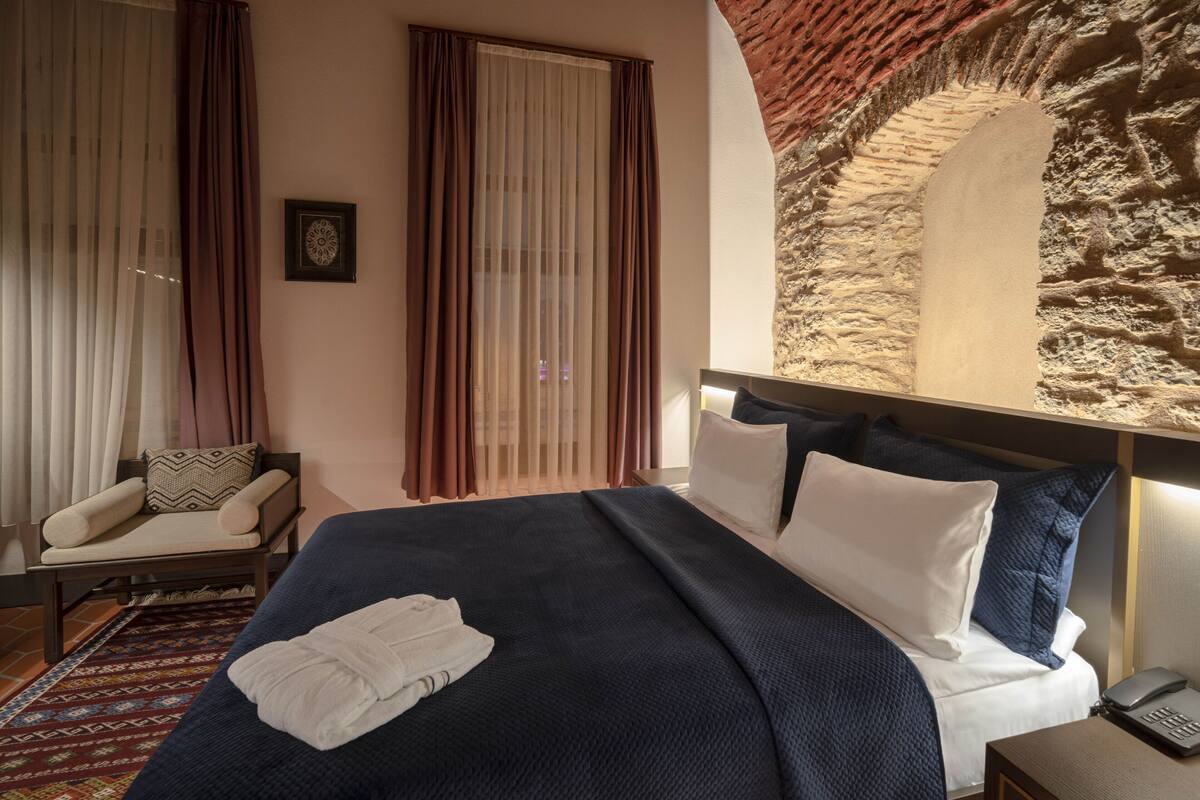A hotel room with a large bed, a chair, a patterned rug, and a stone wall feature.