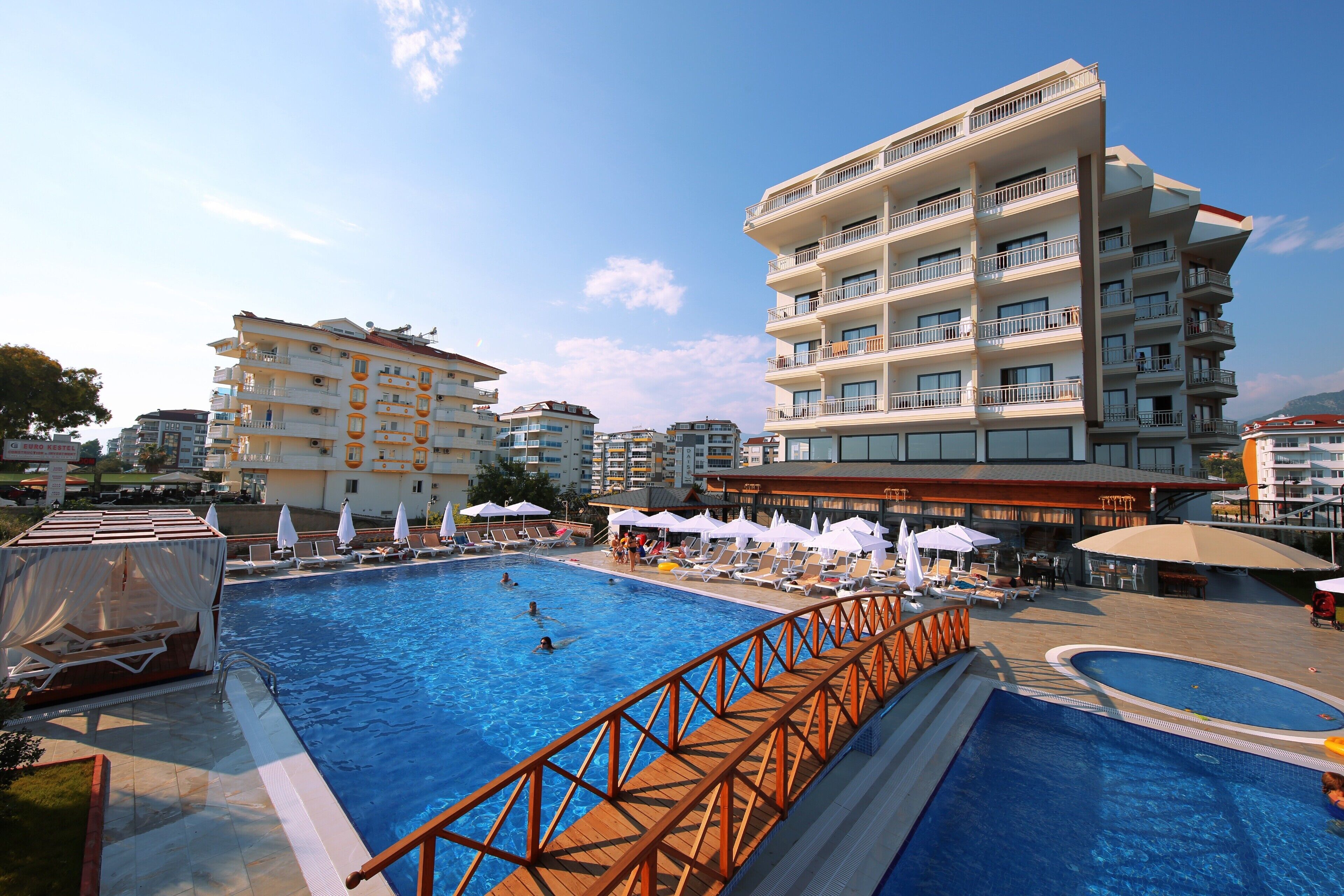 A large outdoor swimming pool with a wooden deck, surrounded by lounge chairs and umbrellas, and adjacent to a multi-story hotel building.