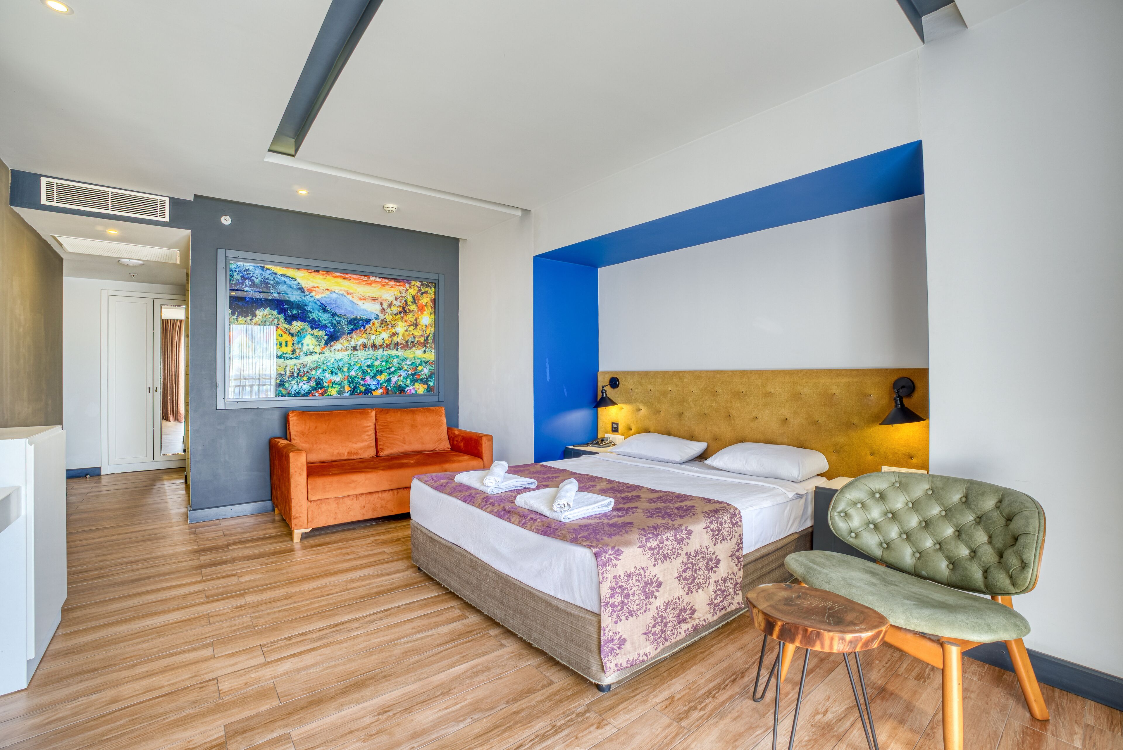 A modern hotel room with a large bed, an orange sofa, a green armchair, and a painting on the wall.