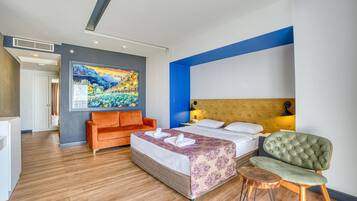A modern hotel room with a large bed, an orange sofa, a green armchair, and a painting on the wall.