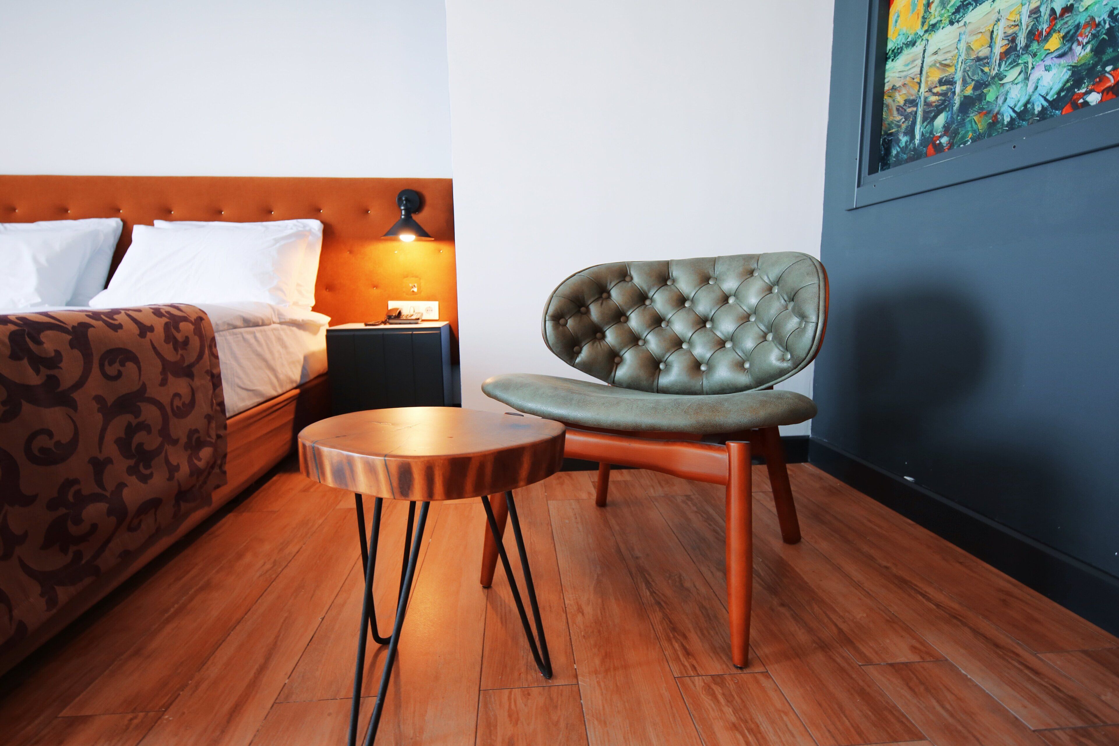 A hotel room with a bed, a chair, a small table, and a nightstand with a lamp.