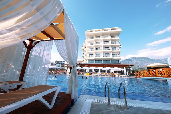 A swimming pool area with lounge chairs and umbrellas, a poolside deck, and a multi-story building in the background. - SEY BEACH HOTEL &SPA (Alanya)