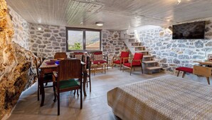 Une pièce rustique aux murs de pierre, avec une table à manger en bois, des chaises rouges, un lit et un téléviseur fixé au mur.