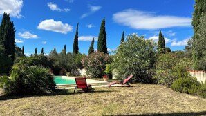 Un patio trasero con piscina, dos sillones reclinables rojos y una variedad de árboles y arbustos.