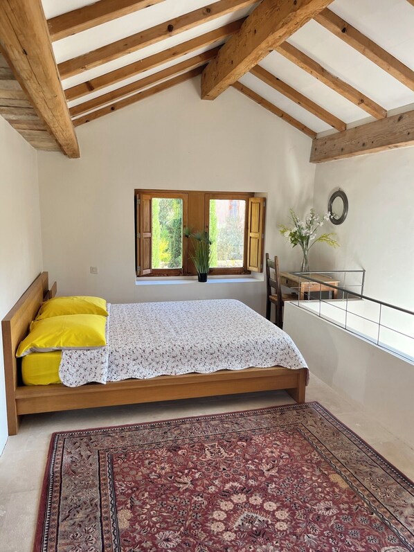 Una camera da letto con un letto in legno, un tappeto rosso con motivi decorativi, una finestra vista su del verde e un tavolino con un vaso di fiori.