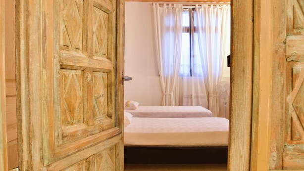 Una puerta de madera abierta que da a una habitación con una cama, una ventana con cortinas y un suelo de baldosas.
