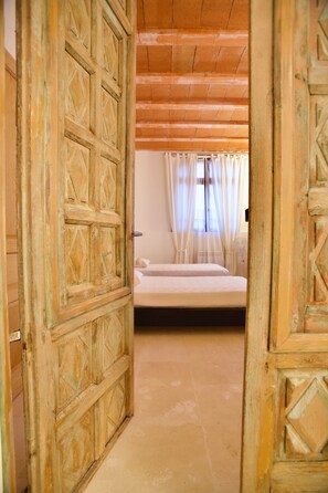 Una porta in legno aperta che dà su una stanza con un letto, una finestra con tende e un pavimento piastrellato.