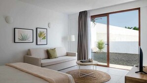 A modern hotel room with a sofa, a small round table, a TV, and a large sliding glass door leading to a balcony with a view of a white building and a palm tree. - Villa Amade, con Piscina Privada (Playa Blanca (Yaiza))
