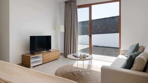 Ein modernes Hotelzimmer mit einem Flachbildfernseher auf einem Holzständer, einem kleinen runden Tisch mit Büchern und einer Lampe, einer Couch und einer großen Schiebeglasdoor zu einem Außenbereich.
