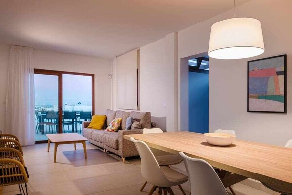 A modern living room with a sofa, dining table, and chairs. There is a large window and a door leading to a balcony. - Villa Maialen, con Piscina Privada (Playa Blanca (Yaiza))
