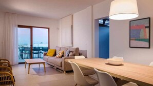 A modern living room with a sofa, dining table, and chairs. There is a large window with curtains, a ceiling light, and a painting on the wall. - Villa Alba, con Piscina Privada (Playa Blanca (Yaiza))
