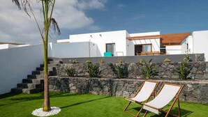 Uma villa moderna com um muro de pedra, uma palmeira e duas espreguiçadeiras em um gramado bem cuidado.