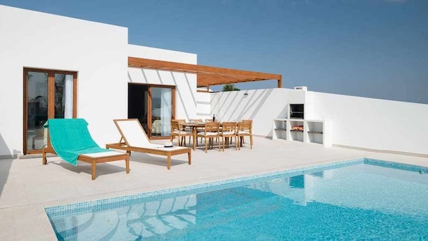 Uma área moderna de piscina ao ar livre, com piscina azul límpida, espreguiçadeiras de madeira e uma mesa de jantar com cadeiras.