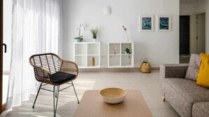 Un salon moderne avec un fauteuil en osier, un canapé gris agrémenté d’un coussin jaune, une console blanche ornée de plantes et des œuvres d’art encadrées accrochées au mur.