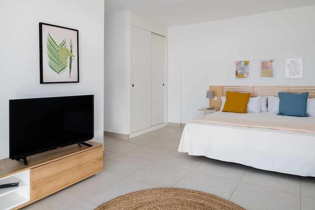 Ein modernes Hotelzimmer mit einem Flachbildfernseher auf einem Holzboden, einem Bett mit weißer Bettwäsche und bunten Kissen, einem weißen Kleiderschrank und einer gerahmten Blattform-Kunstwerk an der Wand.