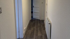 En hall med trägolv, vita väggar och en taklampa. Det finns två dörrar på vänster sida och en på höger sida.