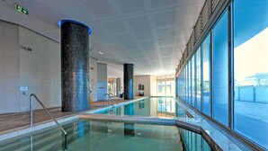 A swimming pool with glass walls and metal railings, surrounded by a corridor with a tiled floor and a high ceiling.