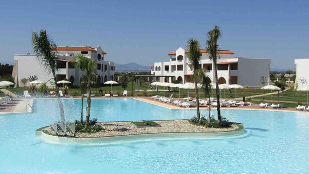 Ett semesterparadis med en stor swimmingpool, palmer och vita byggnader med röda tak.