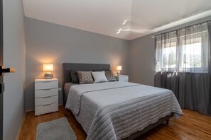 A bedroom with a bed, two bedside tables, a nightstand, a window with curtains, and a wooden floor.