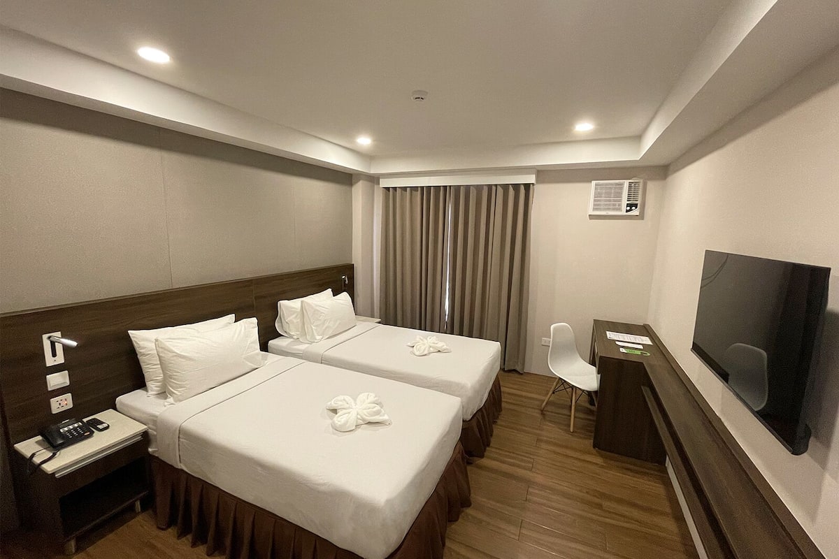 A hotel room with two beds, a desk with a chair, a flat-screen TV, and a window with curtains.